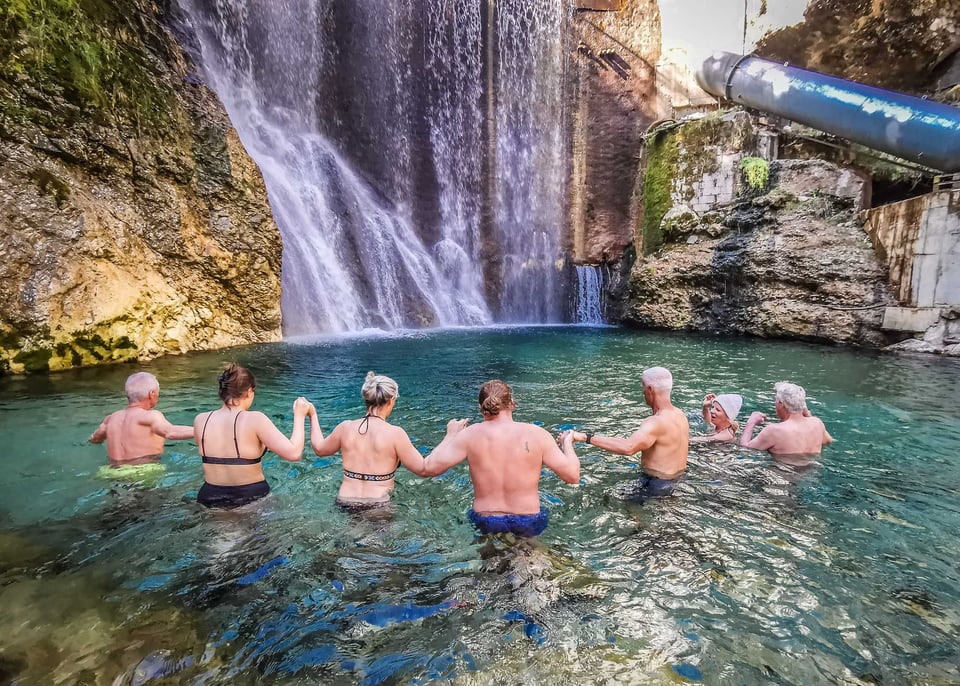 Gruppe badet gemeinsam in einem türkisfarbenen Becken vor einem Wasserfall